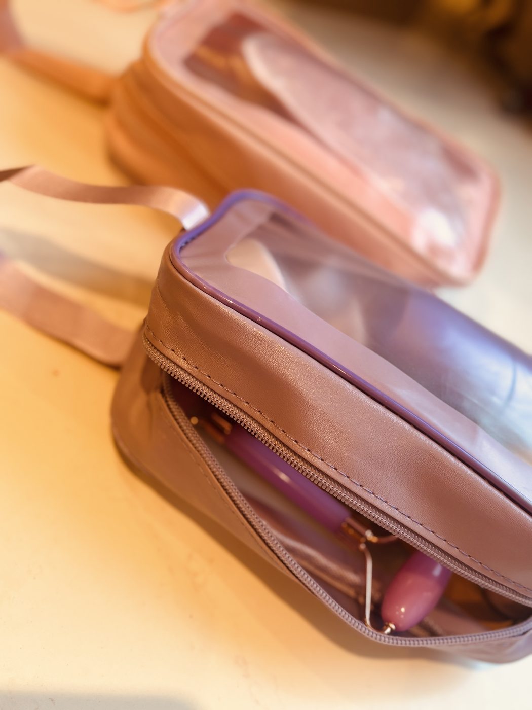 Deux petites trousses de toilette zippées à fenêtre transparente, l'une rose poudré, l'autre lilas, posées sur un plan clair