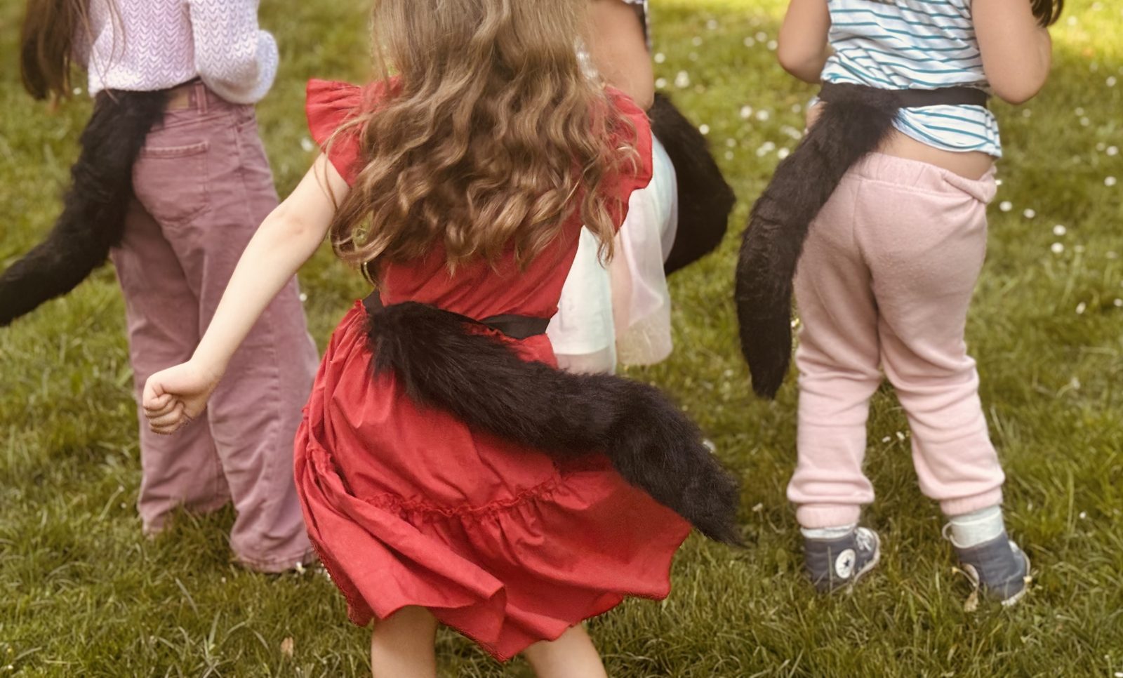 Des enfants de dos avec une queue de chat en fourrure accrochée à la ceinture, en plein jeu dans le jardin