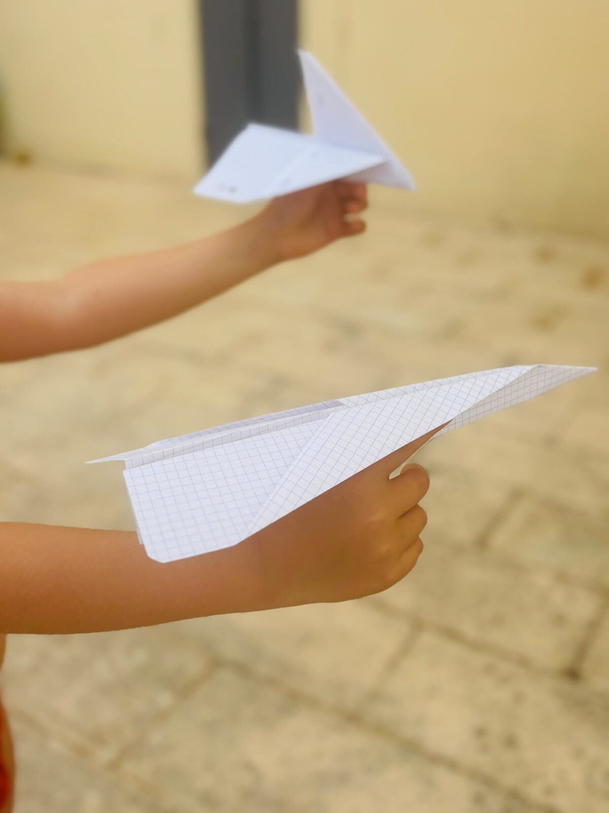 Deux bras d'enfants tendus, chacun prêt à lancer son avion en papier, l'un en papier quadrillé