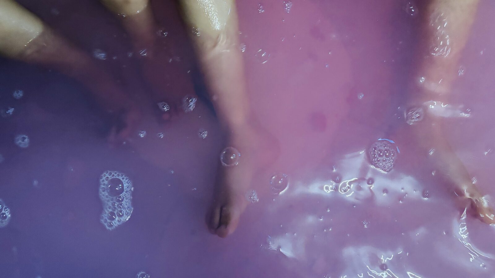 Eau de bain teintée en dégradé violet et rose avec des bulles, pieds d'enfants dans l'eau