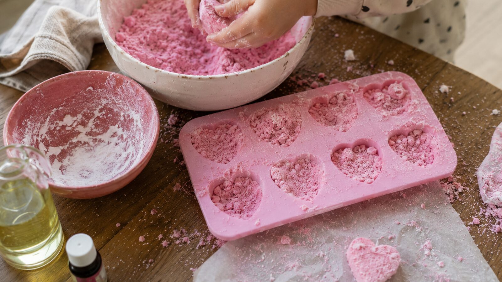 Mains d'enfant tassant la préparation rose dans un moule en silicone en forme de cœurs, à côté d'un saladier de poudre, d'un flacon d'huile et d'un petit flacon de colorant