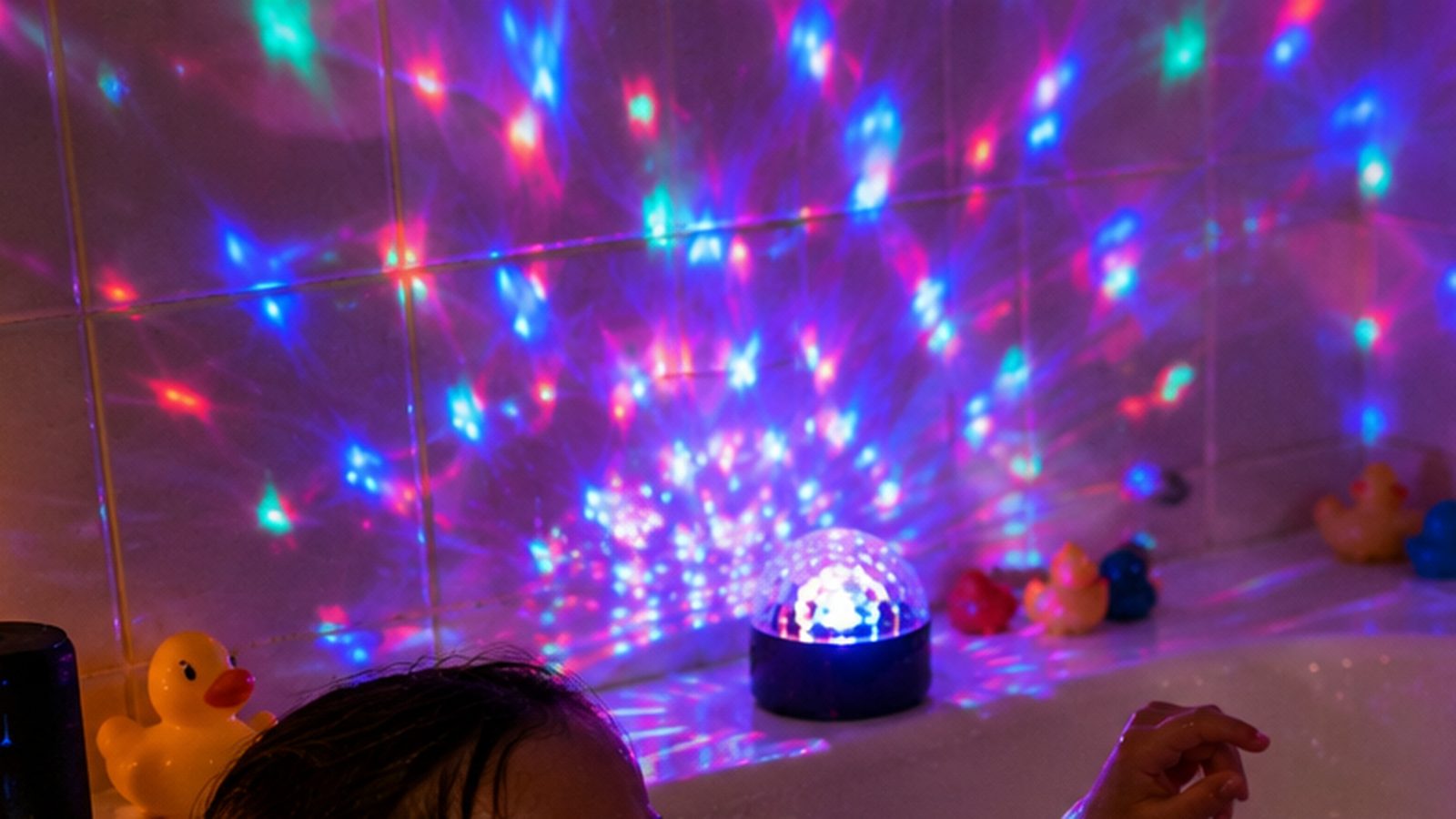 Une boule disco à piles posée sur le rebord de la baignoire projette des points lumineux colorés sur le carrelage, entourée de canards en plastique