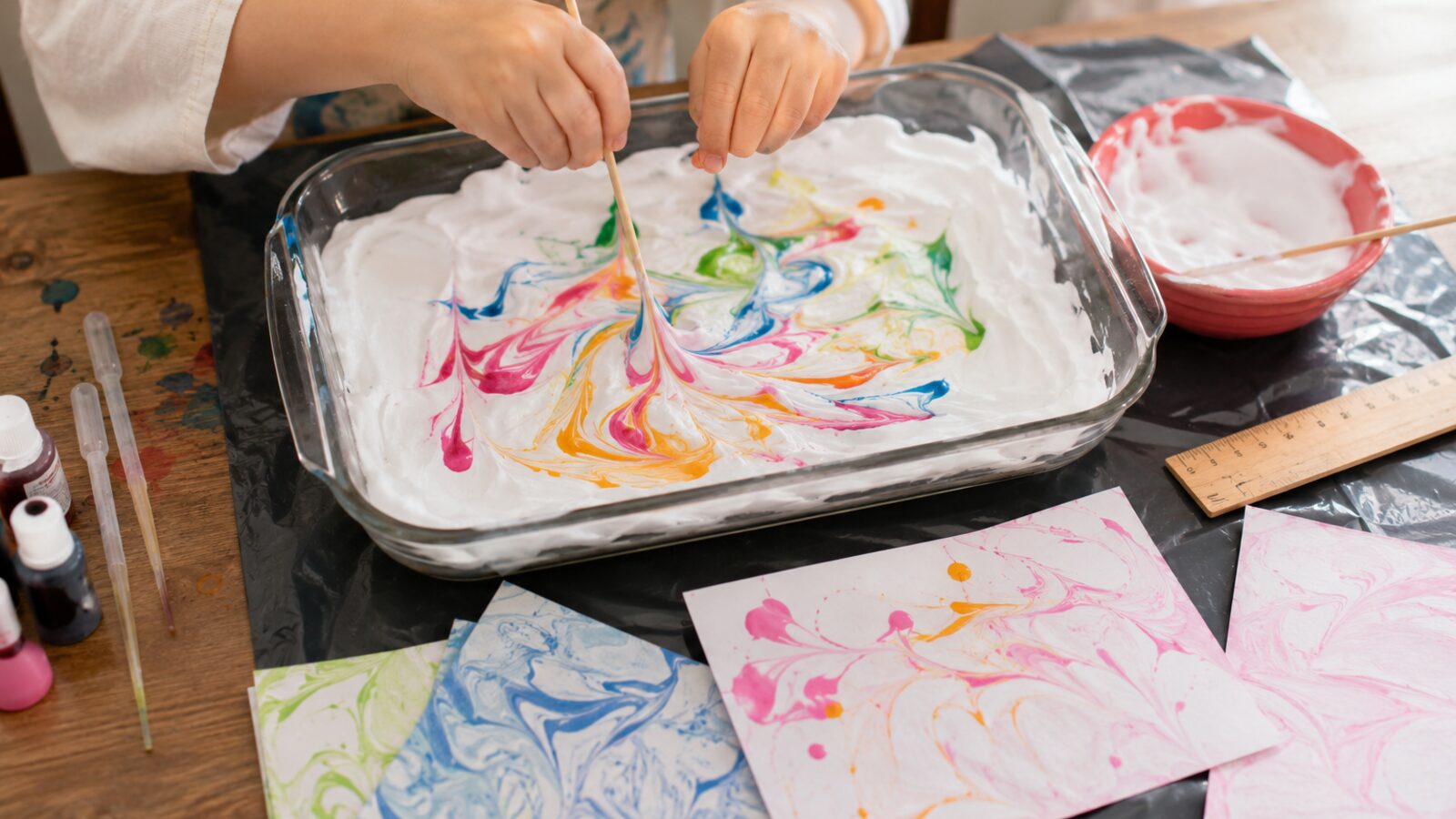 Un enfant trace des tourbillons colorés à la pique en bois dans un plat de mousse à raser, avec des cartes marbrées qui sèchent au premier plan