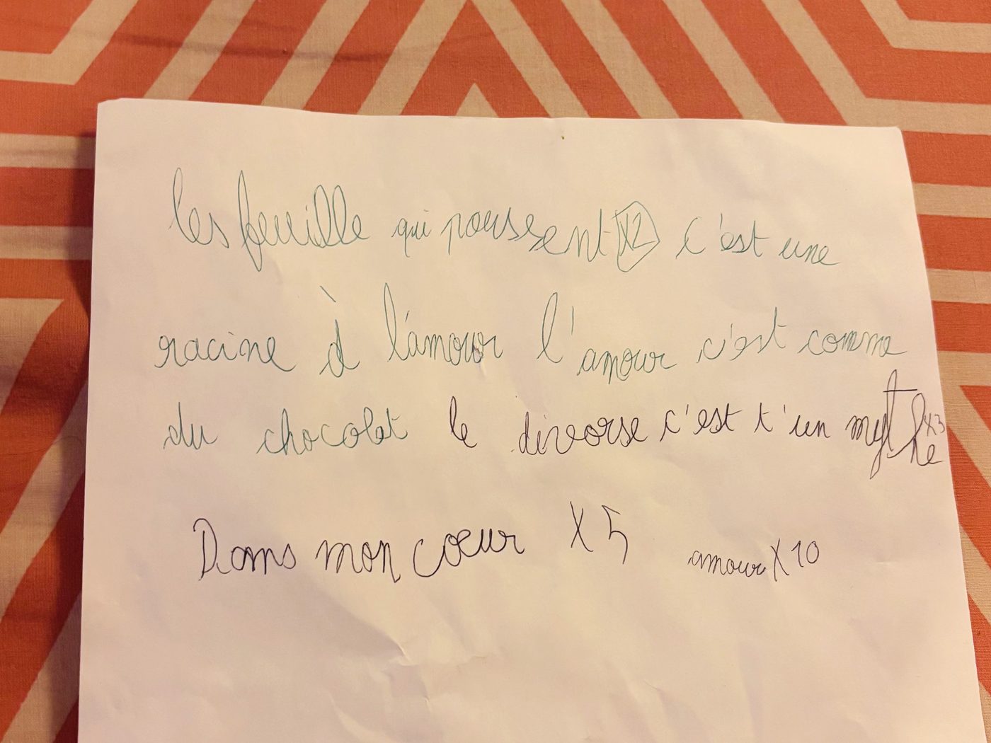 Une feuille blanche posée sur une nappe orange, couverte de paroles de chanson écrites en cursive par deux enfants, l'un au stylo vert et l'autre au stylo bleu, avec des indications de répétition notées en bout de ligne