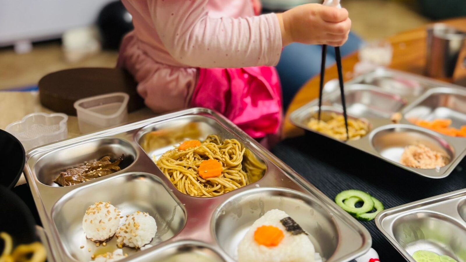 Mains d'enfant tenant des baguettes surmontées d'un petit lapin, au-dessus d'un plateau à compartiments garni de nouilles décorées d'une fleur de carotte, de boulettes de riz au sésame, d'un onigiri triangulaire à l'algue nori et de fleurs de concombre