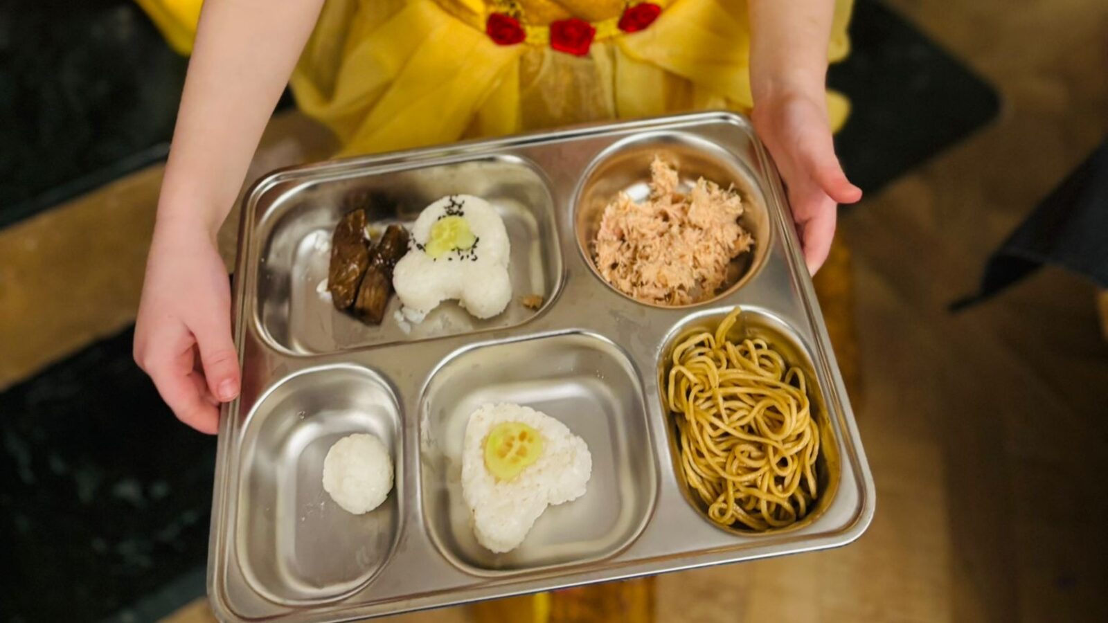 Enfant en robe jaune tenant à deux mains un plateau à compartiments en inox garni de boulettes de riz en forme de cœur et d'animal, de thon émietté et de nouilles