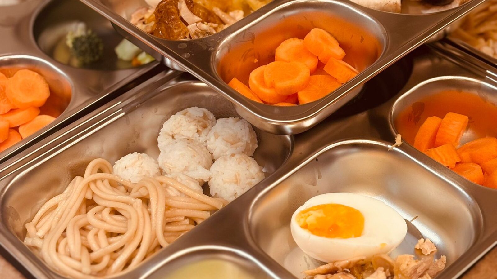 Plateaux à compartiments en inox garnis de nouilles, rondelles de carotte, boules de riz, demi-œufs, concombre et champignons, façon bento du quotidien