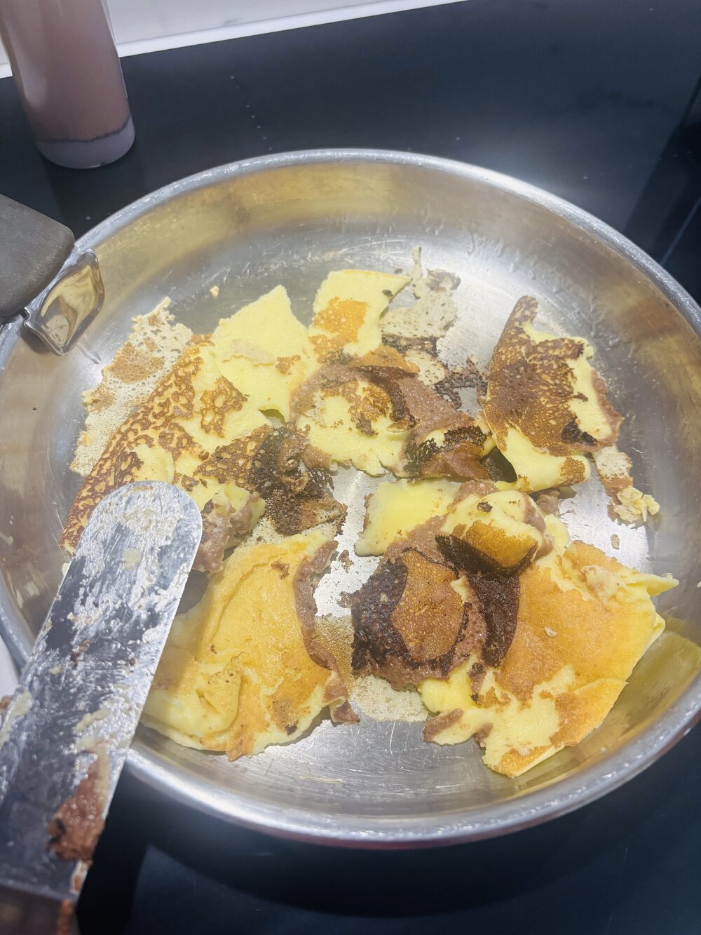 Une première crêpe marbrée déchirée en morceaux dans la poêle, marbrures de chocolat inégales et un peu brûlées, spatule posée à côté