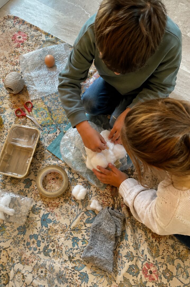 Deux enfants emballent soigneusement un œuf dans du coton et du papier bulle, entourés de ficelle, ruban adhésif et barquettes de récup