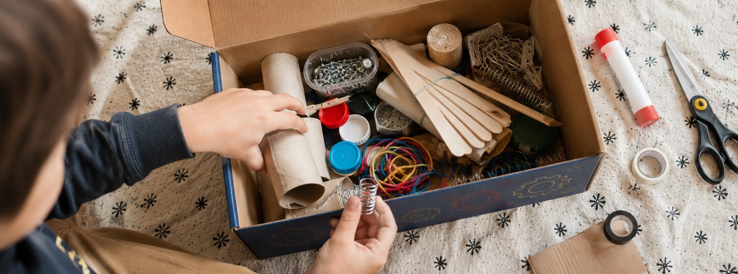 Un enfant fouille dans une boîte remplie de rouleaux en carton, bâtonnets, élastiques, bouchons, ressorts et trombones