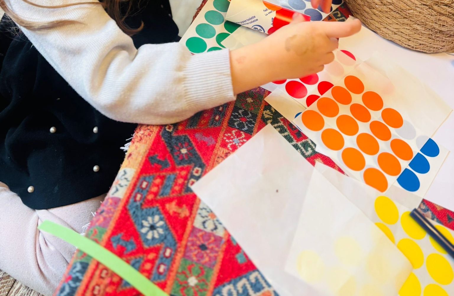Main d'enfant qui décolle une grosse gommette orange d'une planche pour la coller, à côté d'autres planches de gommettes vertes, bleues et jaunes posées sur un tapis coloré