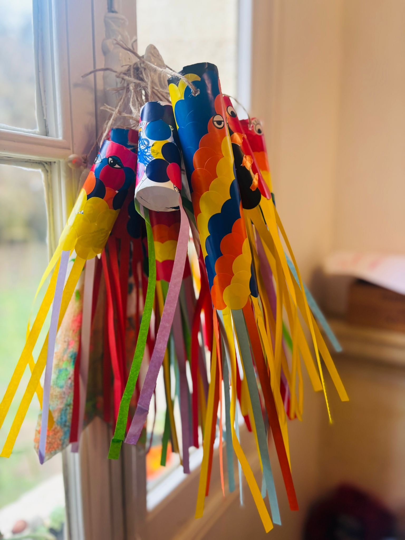 Plusieurs koinobori en rouleaux couverts de gommettes colorées, accrochés à une ficelle devant une fenêtre, leurs longues queues en papier multicolore qui pendent dans la lumière du jour