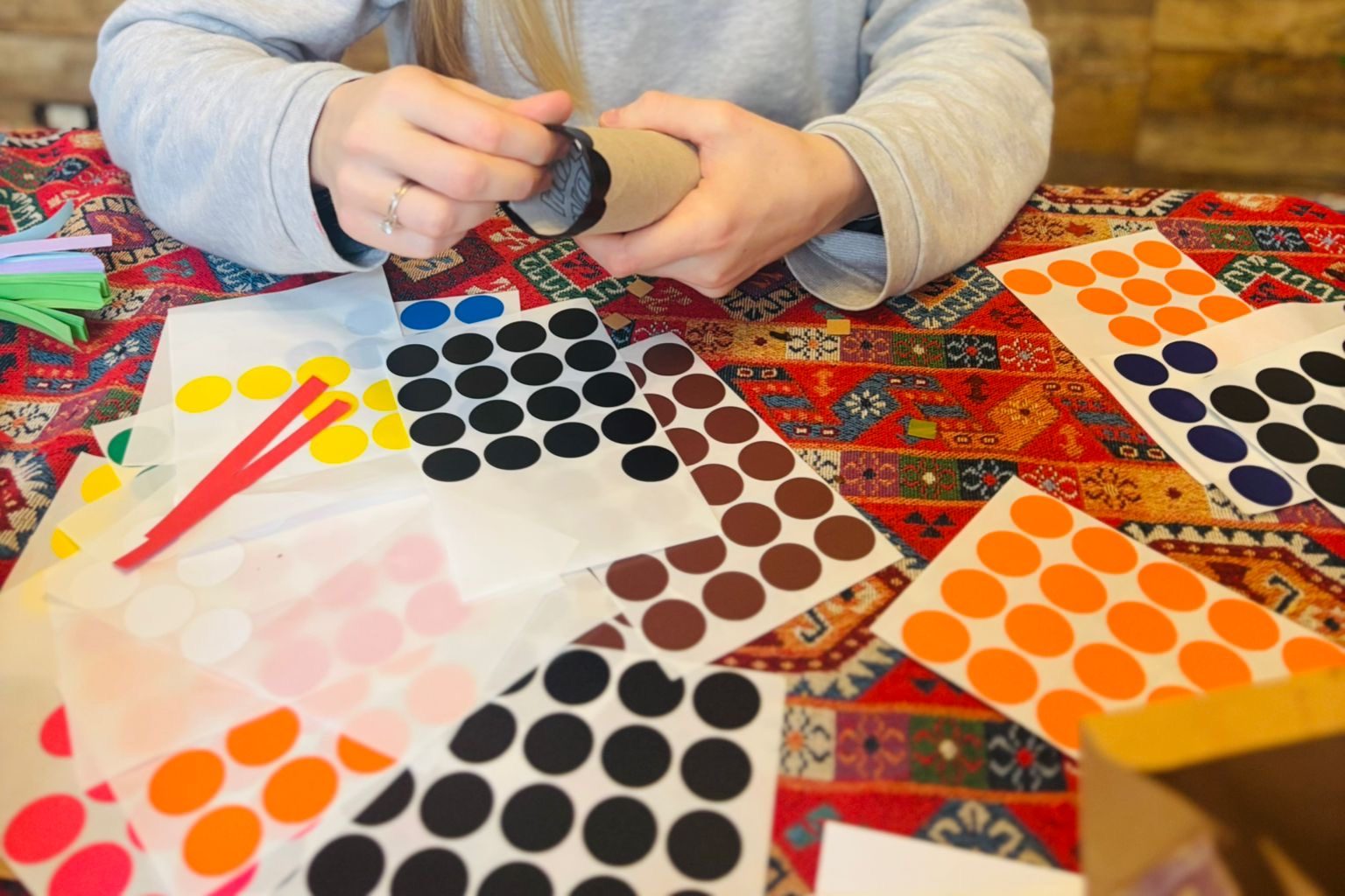 Des mains d'adulte qui entourent un rouleau de carton, posées au-dessus d'une table couverte de planches de grosses gommettes rondes de toutes les couleurs et de fines bandes de papier rouge