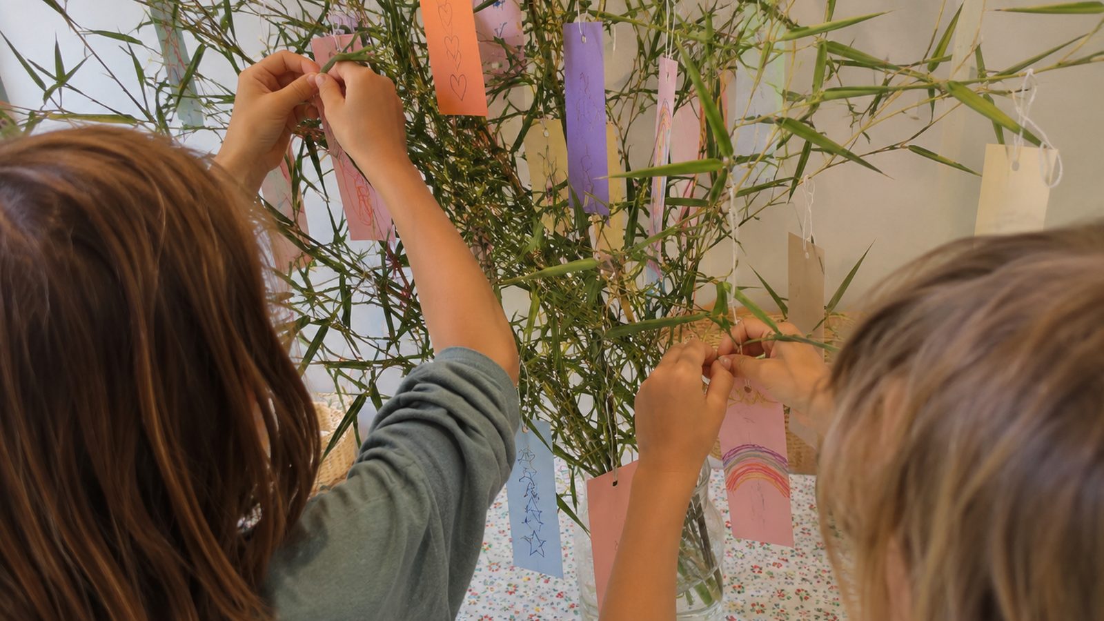 Deux enfants vus de dos accrochent leurs étiquettes de souhaits colorées aux branches d'un bambou, certaines décorées de dessins d'enfant comme un arc-en-ciel, des étoiles et des cœurs