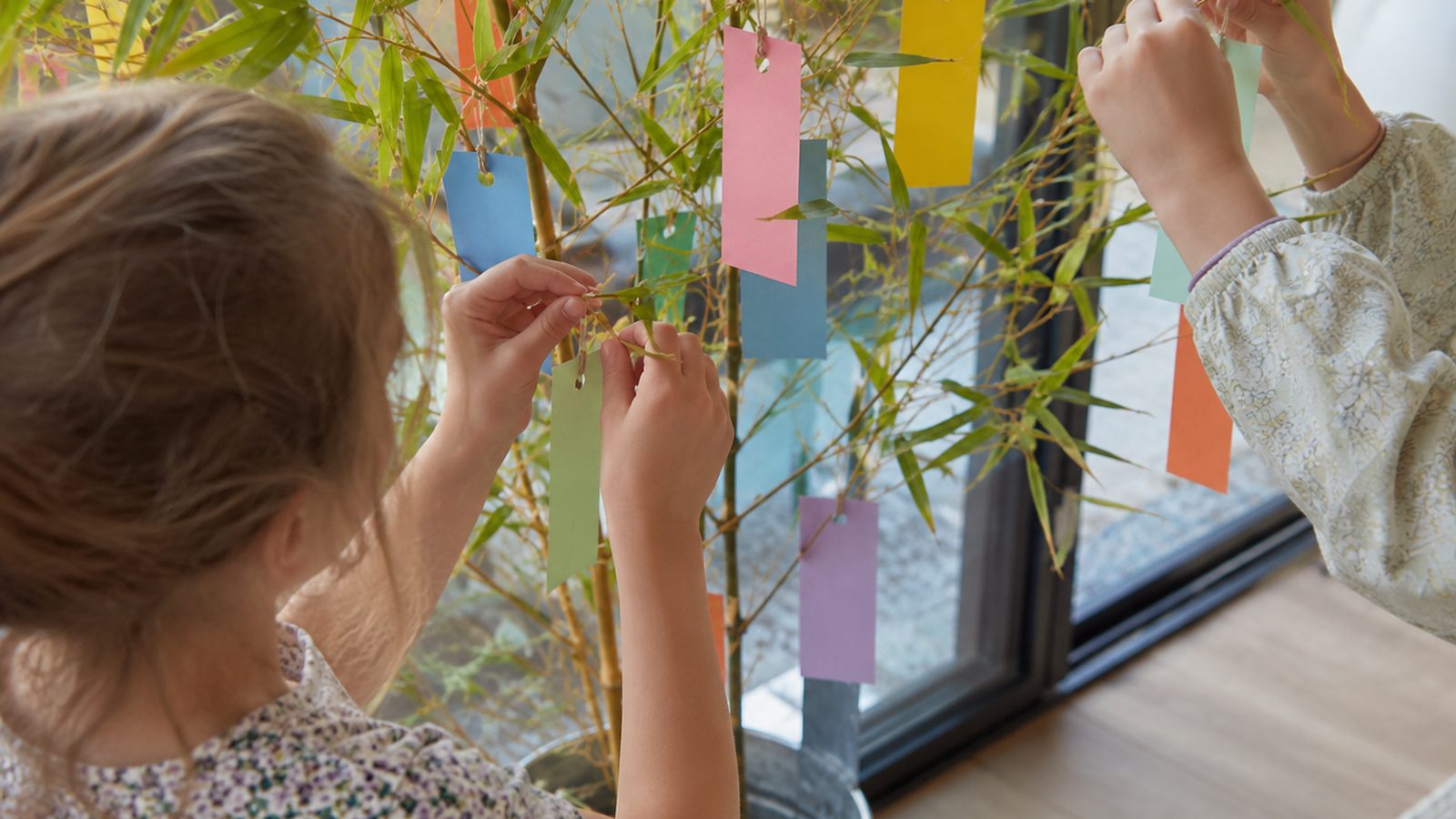 Un adulte et un enfant, vus de dos, accrochent ensemble des étiquettes de couleur aux branches d'un bambou près d'une fenêtre