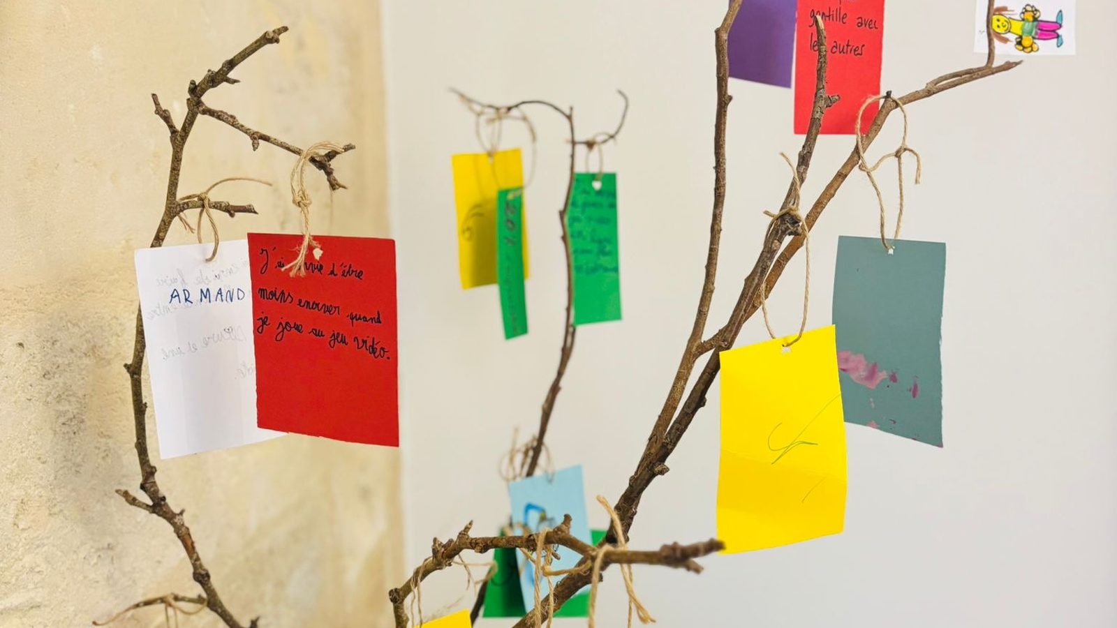 Un arbre à souhaits fait maison : une branche installée dans un vase en métal doré, dans un coin de salon, avec des étiquettes de papier de couleur portant des souhaits manuscrits et des dessins d'enfants