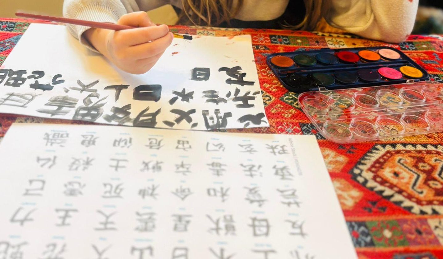 Une main d'enfant, pinceau tenu presque droit, recopie de grands caracteres chinois noirs sur une feuille blanche, avec la planche modèle et la boîte de peinture posées sur la table