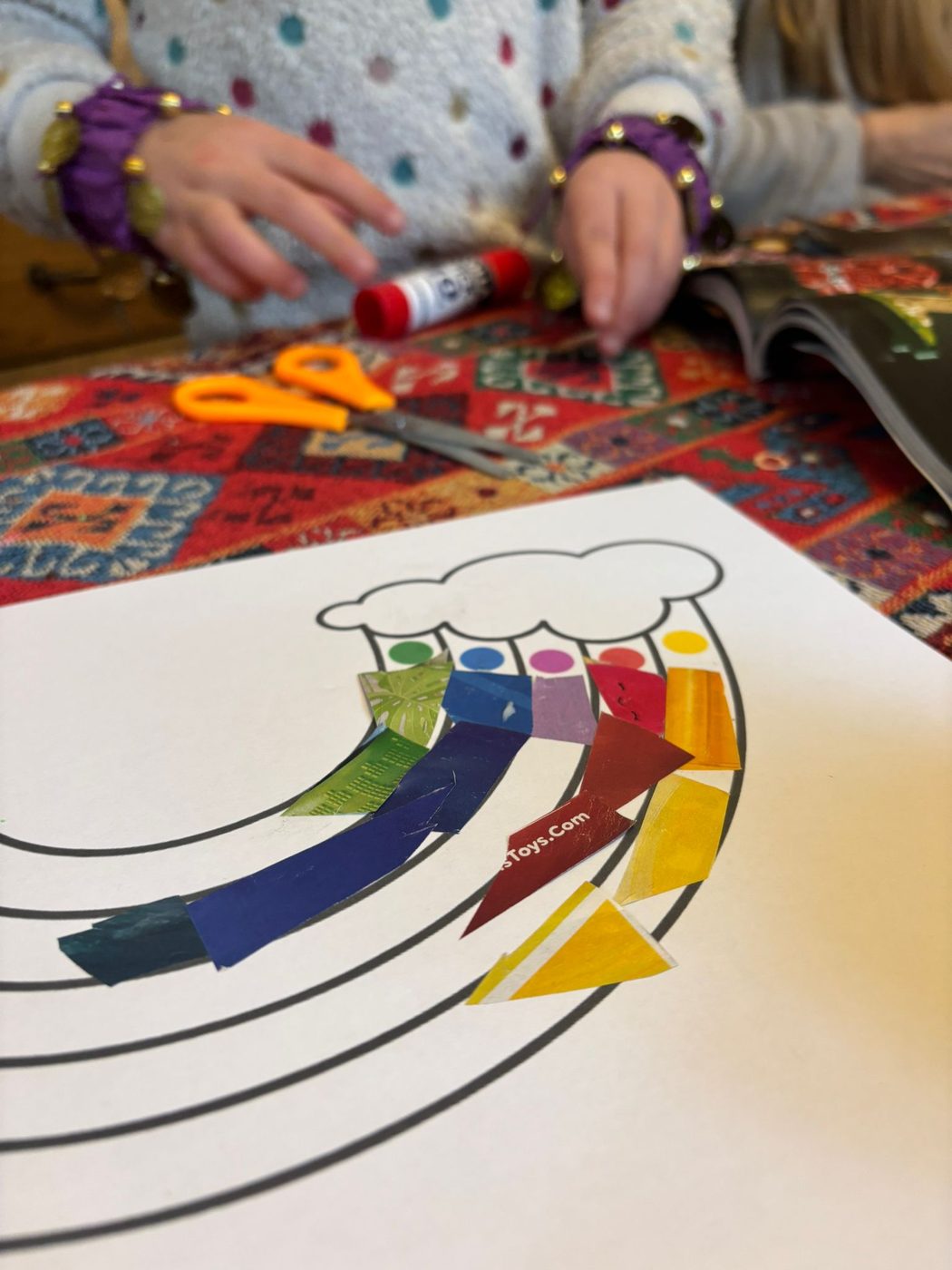 Des mains d'enfant qui collent des bouts de papier découpés dans des magazines sur les bandes d'un arc-en-ciel imprimé, avec des ciseaux et un bâton de colle posés à côté