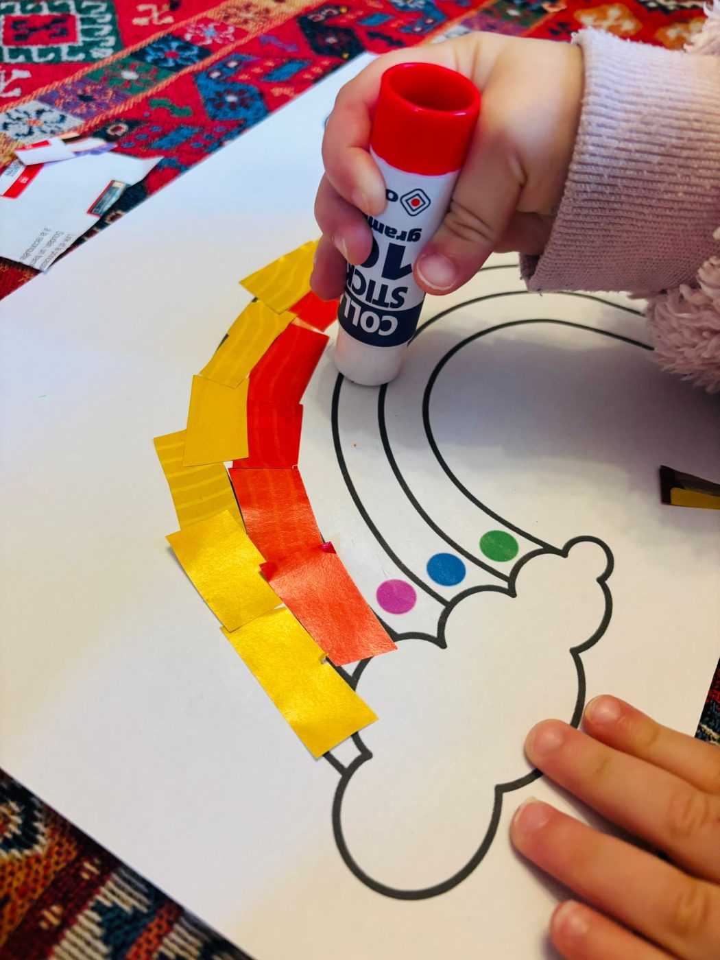 Une main d'enfant qui tient un bâton de colle au-dessus d'un arc-en-ciel dont les bandes chaudes, jaune et orange, sont déjà remplies de morceaux de papier collés