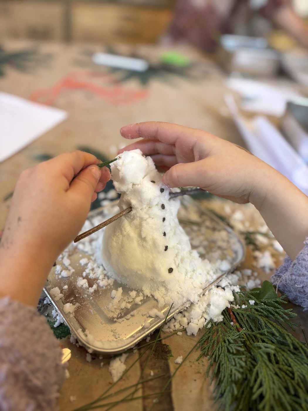 Deux petites mains d'enfant plantent une fine brindille dans le côté d'un bonhomme de neige en bicarbonate, posé sur un plateau métallique saupoudré de neige blanche, avec du thuya à côté