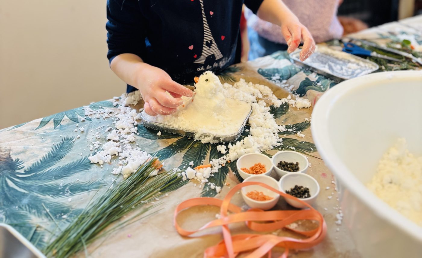 Deux enfants côte à côte décorent leurs bonshommes de neige au bicarbonate sur une table enneigée, avec des bols de lentilles et un ruban corail
