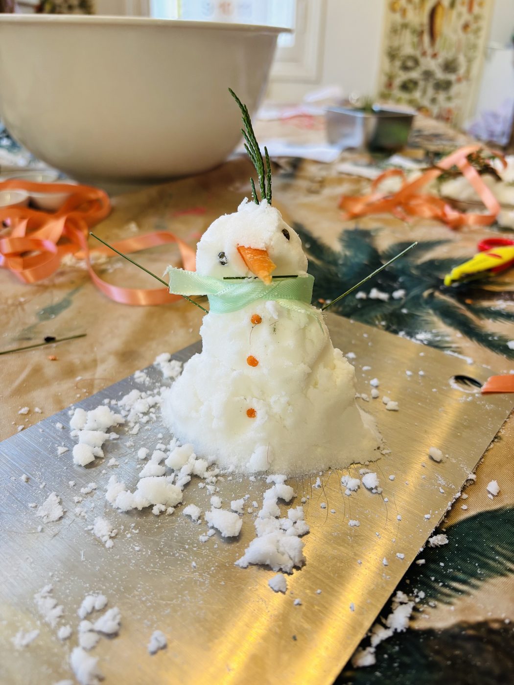 Petit bonhomme de neige en bicarbonate décoré d'un nez en carotte, de lentilles pour les yeux et les boutons, d'un ruban vert pâle en écharpe et d'un brin de thuya sur la tête, posé sur un plateau parsemé de neige