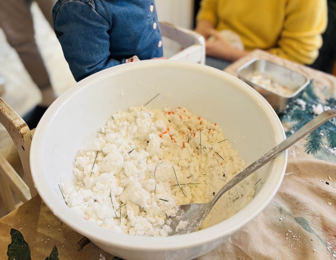 Grand saladier blanc rempli de neige au bicarbonate, blanche et poudreuse avec quelques aiguilles de pin et lentilles orange, une cuillère en métal plantée dedans, sur une table protégée
