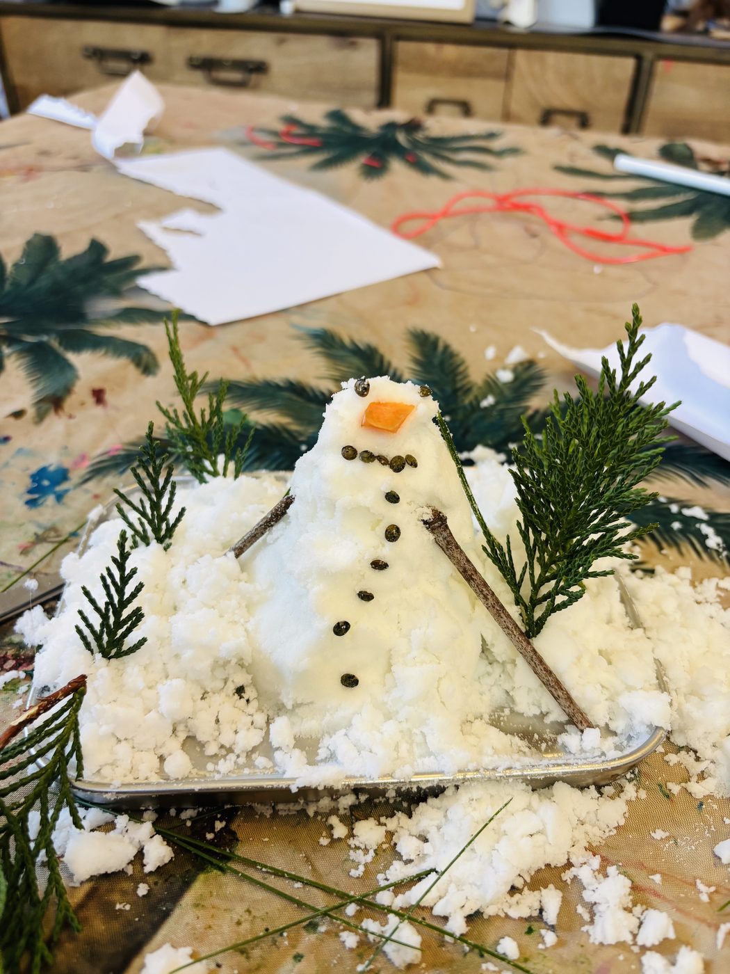 Bonhomme de neige en bicarbonate en forme de petite montagne avec nez en carotte, boutons en lentilles et bras en brindilles, entouré de branchages de thuya dressés comme des sapins sur un plateau enneigé