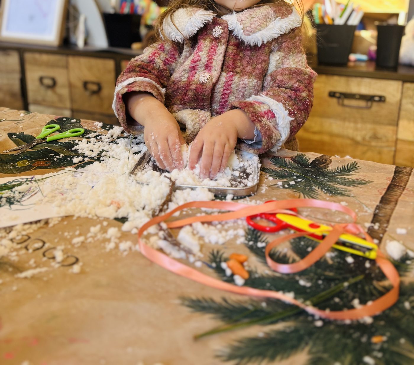Enfant en manteau qui tasse la neige au bicarbonate à deux mains dans une petite barquette en métal, sur une table couverte de neige, avec ruban et ciseaux autour