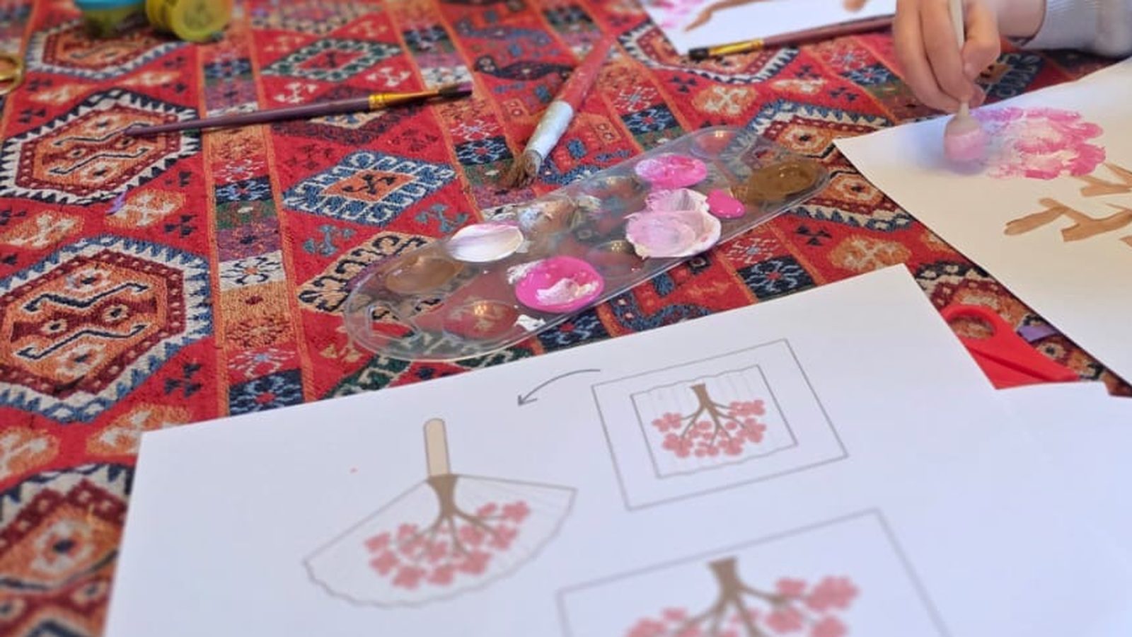 Une palette avec des touches de rose et de blanc à peine mélangées, des pinceaux posés sur une nappe colorée, une main d'enfant qui tamponne une grappe de fleurs roses, et la fiche modèle imprimée à côté