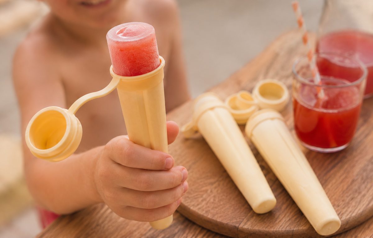 Main d'enfant tenant une glace à l'eau rose grenadine dans un moule silicone façon Mister Freeze, à côté d'autres moules et d'un verre de sirop, sur une table en bois au soleil