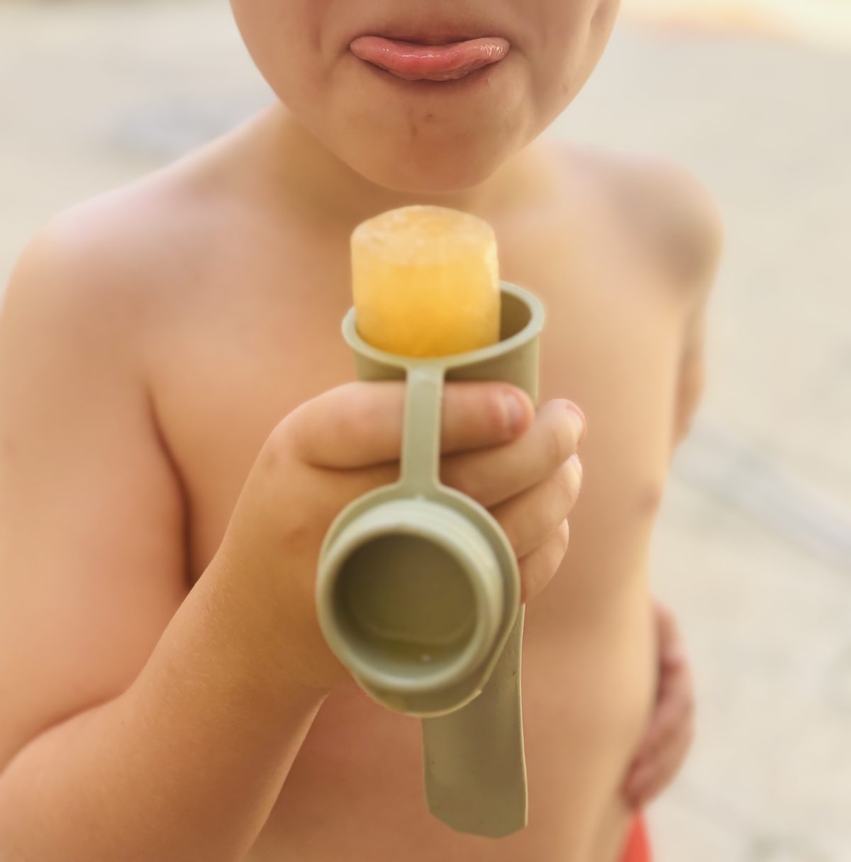 Enfant recadré sous le nez, langue tirée de gourmandise, tenant une glace au jus d'orange dans un moule silicone vert, torse au soleil au bord d'une piscine