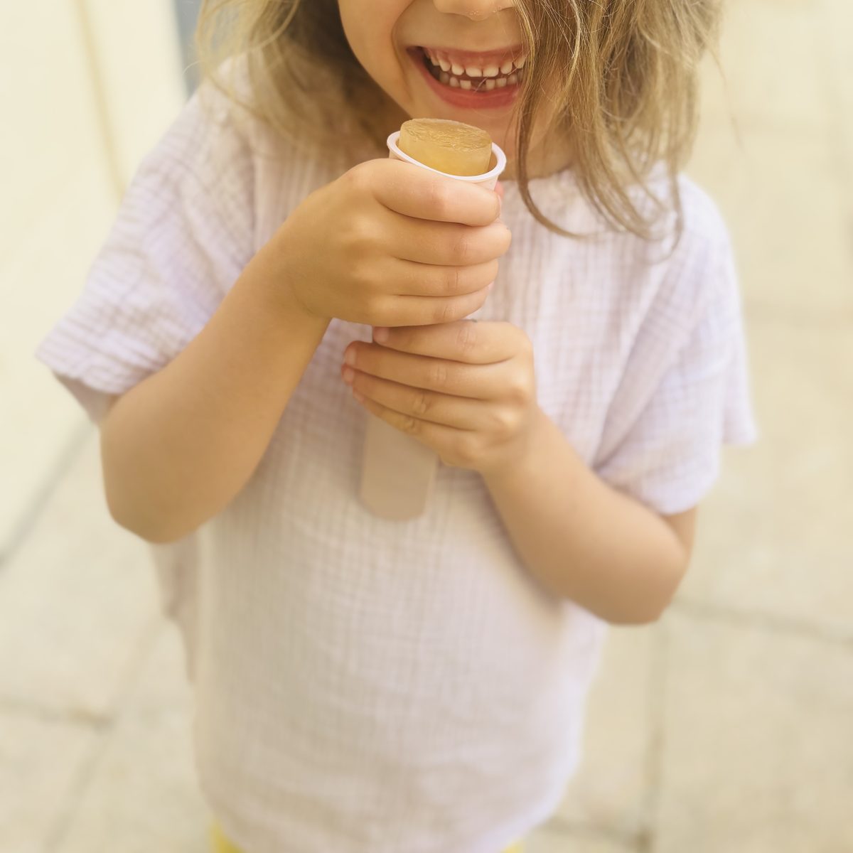 Enfant riant, recadré sous le nez, tenant à deux mains une glace au jus de pomme dans un petit moule pushup blanc, dehors
