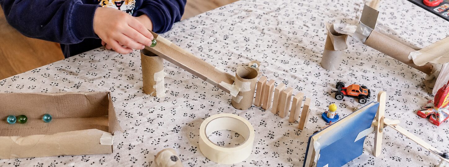 Un enfant teste une rampe en rouleaux de carton avec une bille, à côté de dominos en bois et d'une petite voiture