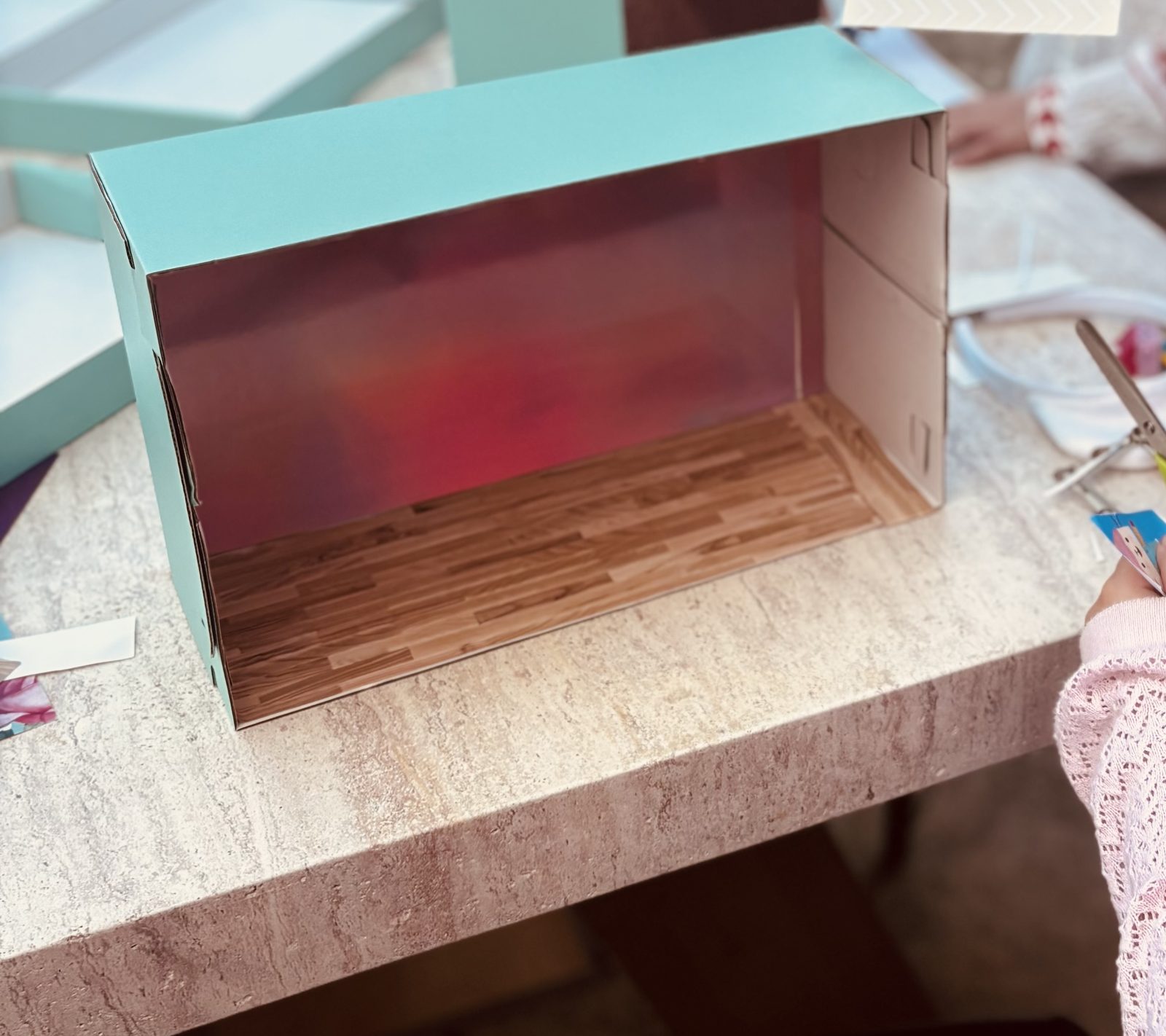 Une enfant tapisse l'intérieur d'une boîte à chaussures transformée en pièce de maison de poupée, avec un sol imitation parquet