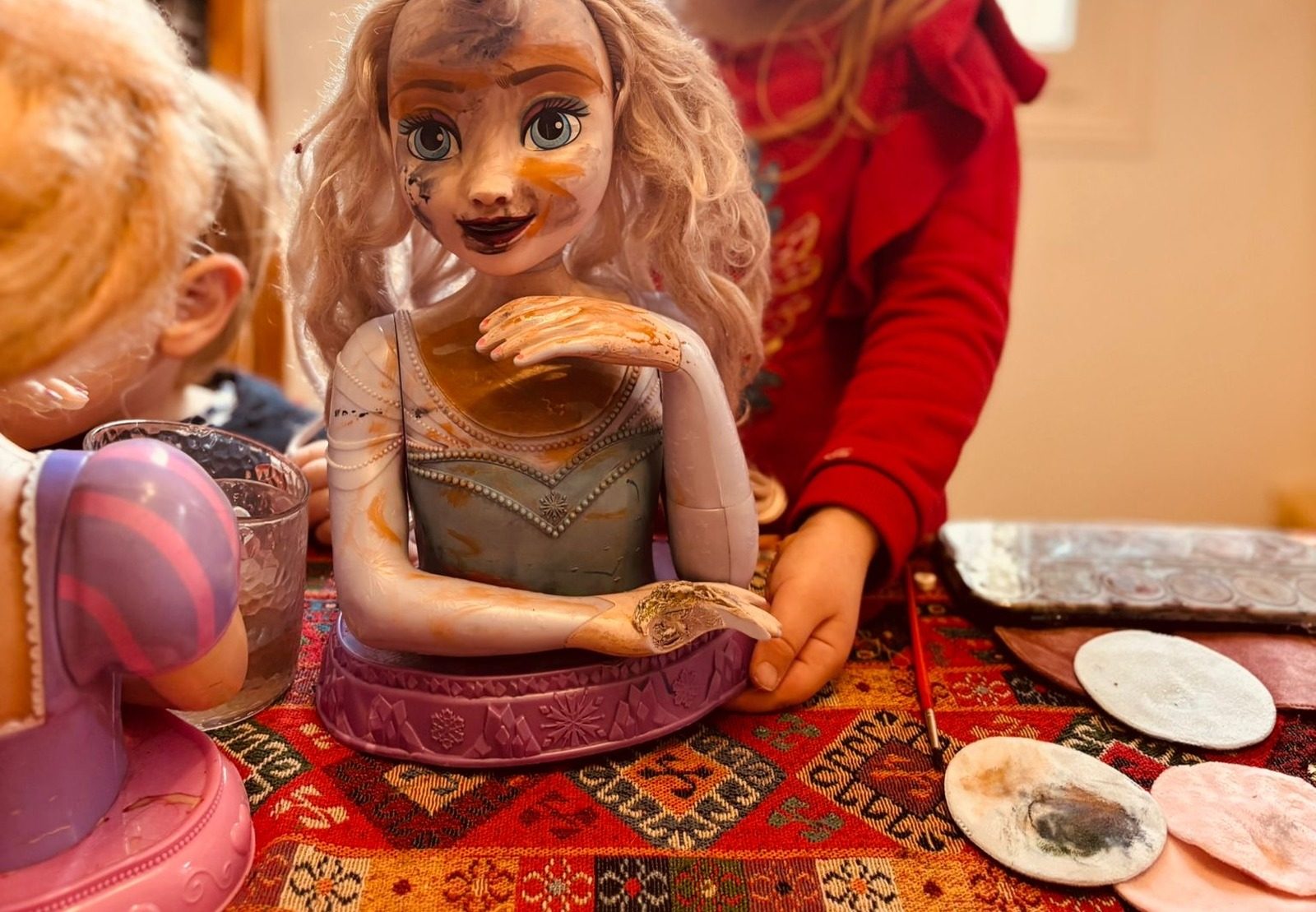 Tête de poupée Reine des neiges couverte de peinture à l'eau, des mains d'enfant qui la tiennent, et à côté des cotons démaquillants tachés de couleur posés sur un tapis