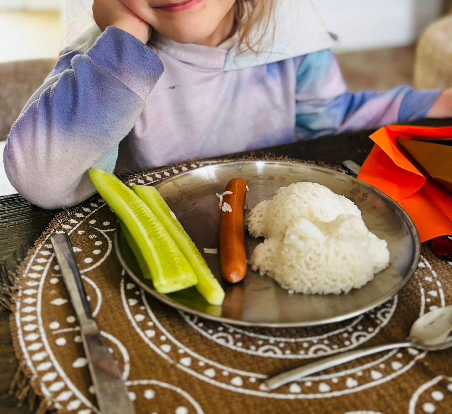 Une assiette avec du riz moulé en forme de chat, une saucisse et des bâtonnets de concombre, une enfant tout sourire derrière