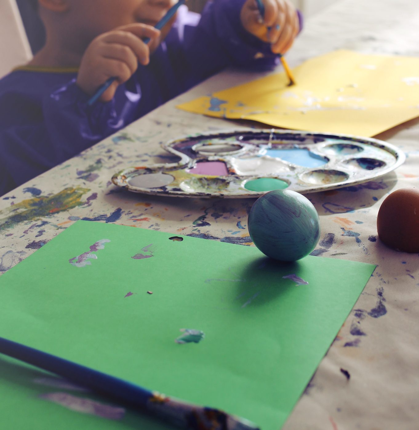 Un enfant peint le fond d'un œuf tenu au bout d'un pinceau, palette de trois couleurs pastel posée sur la table