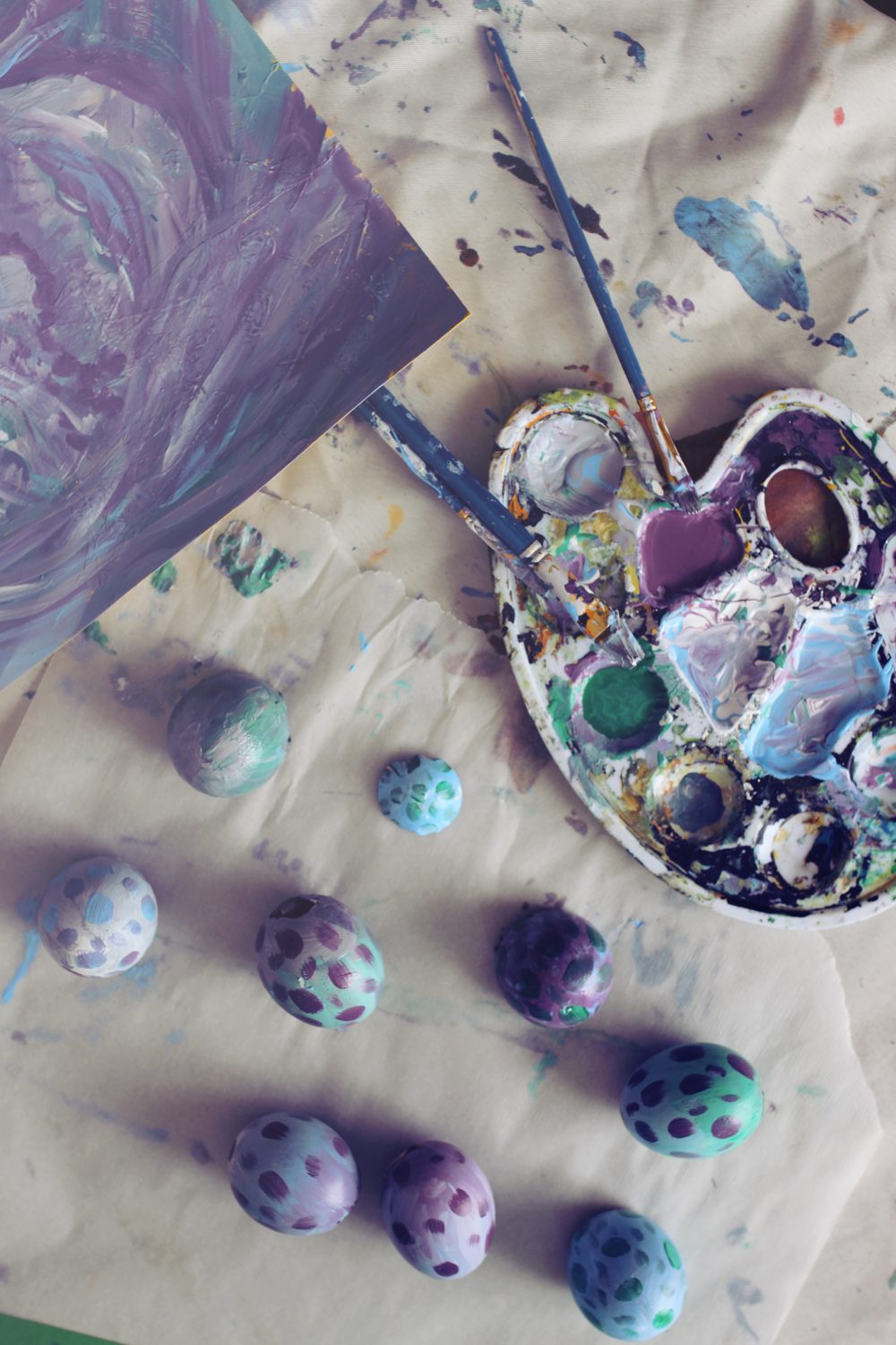 Vue de dessus : palette de peinture avec trois couleurs pastel, œufs mouchetés terminés et pinceaux sur une nappe tachée