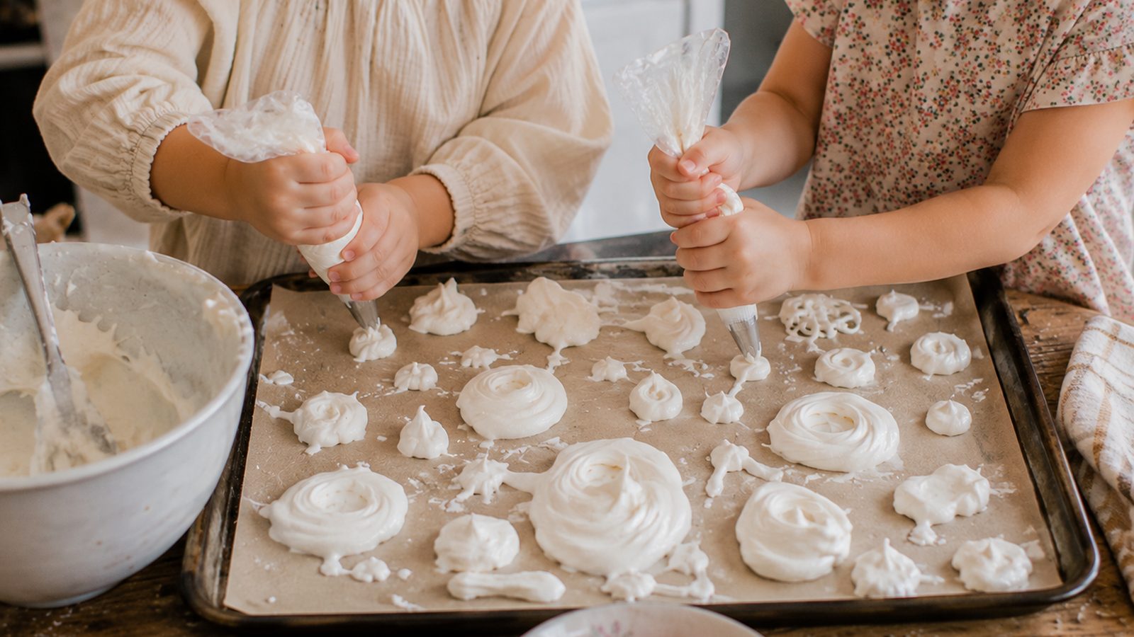 Deux enfants pochent de la meringue en petits nids et coques sur une plaque, poche à douille en main, bols de fruits frais au premier plan