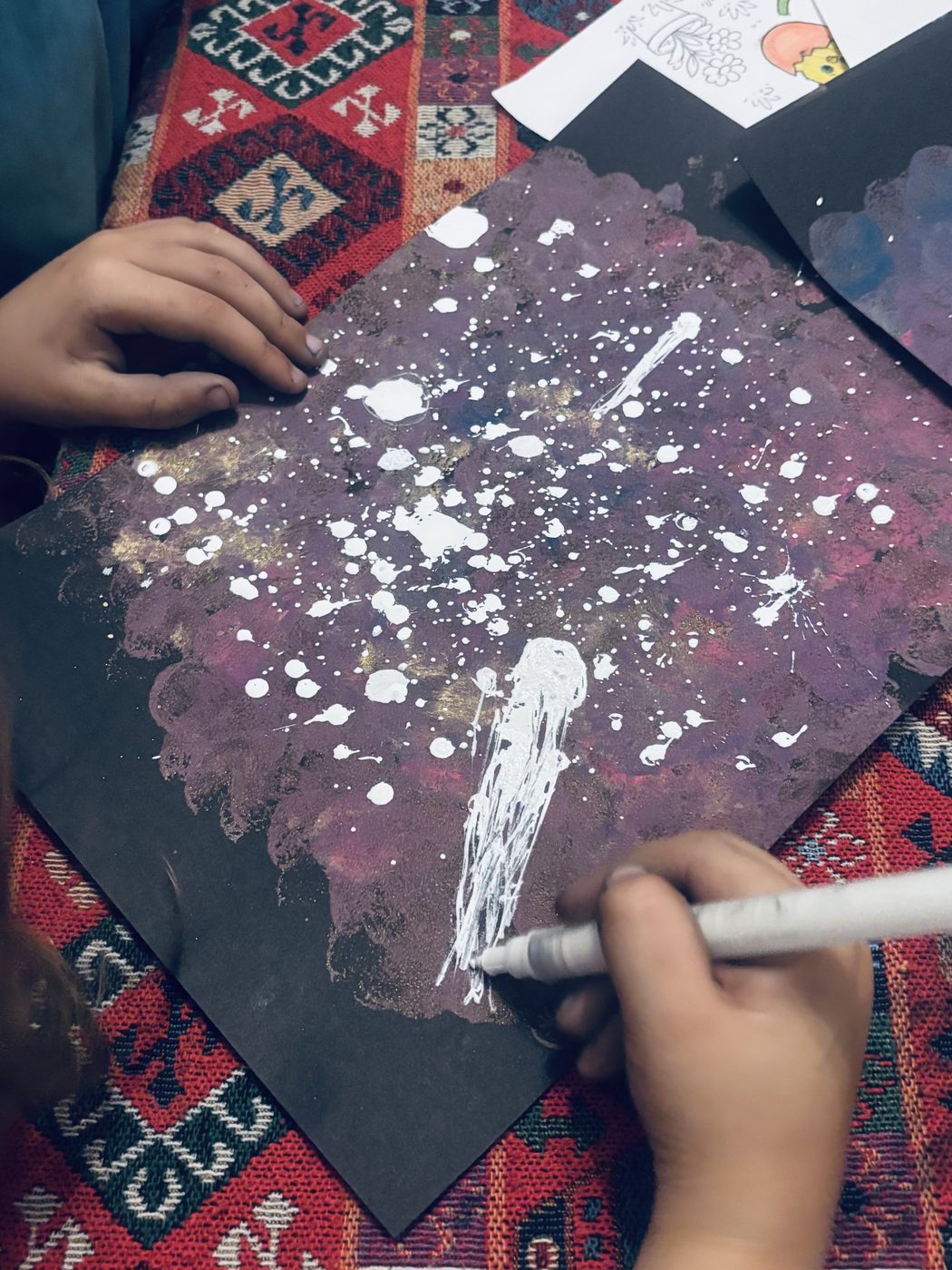 Une galaxie violette couverte de projections de peinture blanche, une main d'enfant la tient pendant qu'une autre ajoute des traits au marqueur blanc