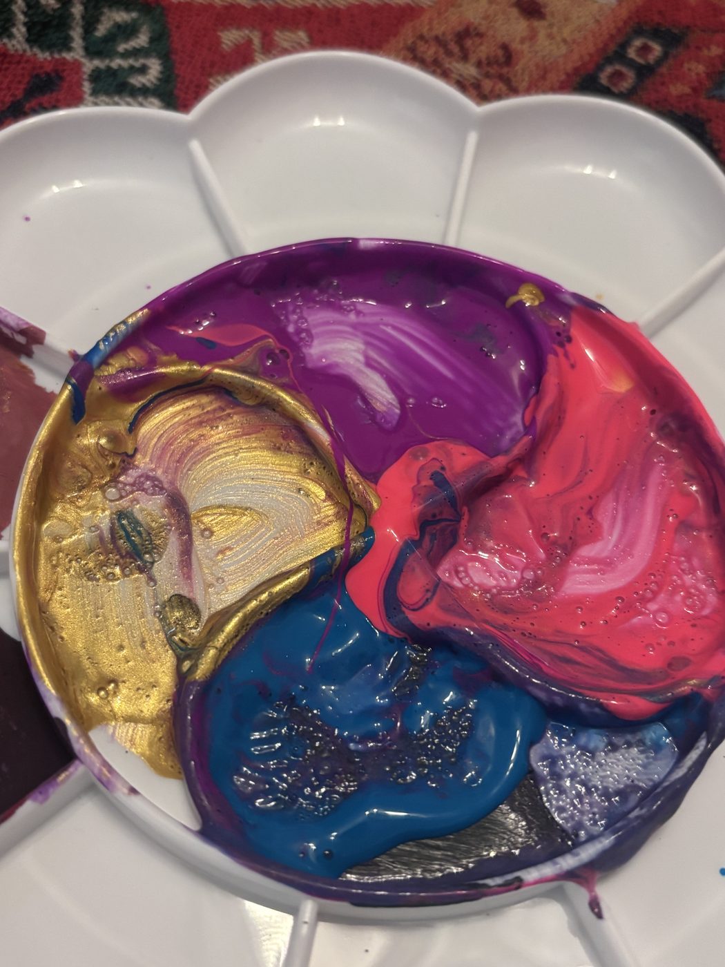 Une palette en plastique blanche remplie de peinture violette, rose fluo, bleue, dorée et argentée, toutes en train de se mélanger