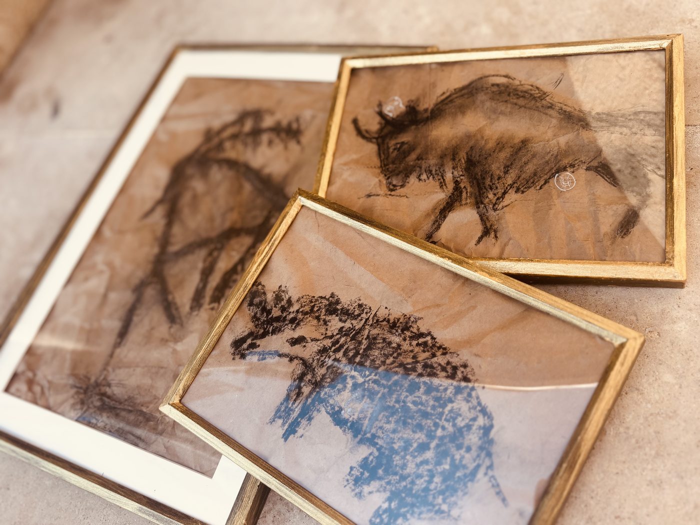 Trois dessins d'animaux au charbon sur papier kraft froissé, encadrés sous verre dans des cadres dorés et blancs posés au sol