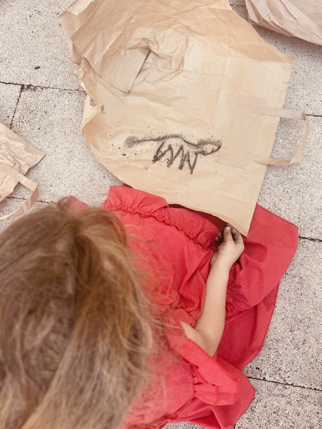 Un enfant vu de dos, assis par terre devant un sac kraft déplié, dessine un cheval au charbon