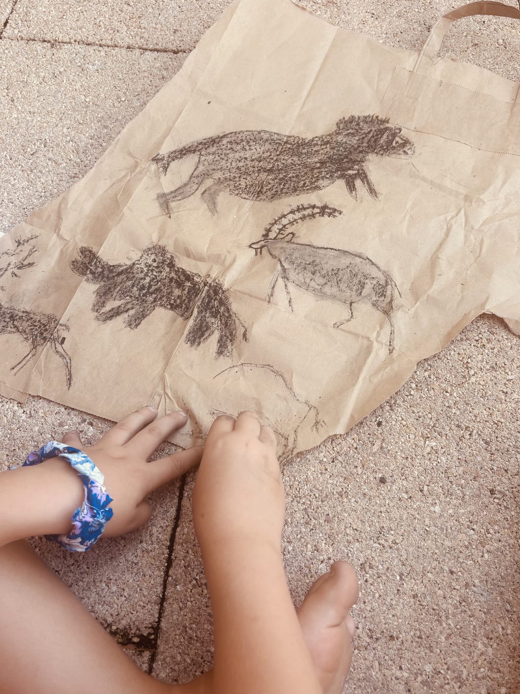 Un sac kraft déplié couvert de dessins au charbon, un lion, un bouquetin, un bison et un cerf, avec des mains d'enfant posées au bord