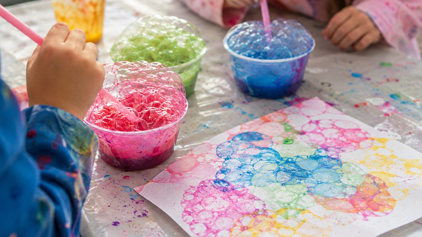 Des enfants soufflent dans des gobelets de peinture à bulles rose, verte et bleue, à côté d'une feuille couverte d'empreintes de bulles multicolores