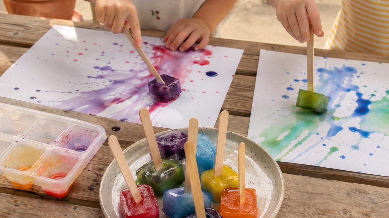 Deux enfants font glisser des glaçons colorés sur des feuilles blanches, créant des traînées violettes, bleues et vertes