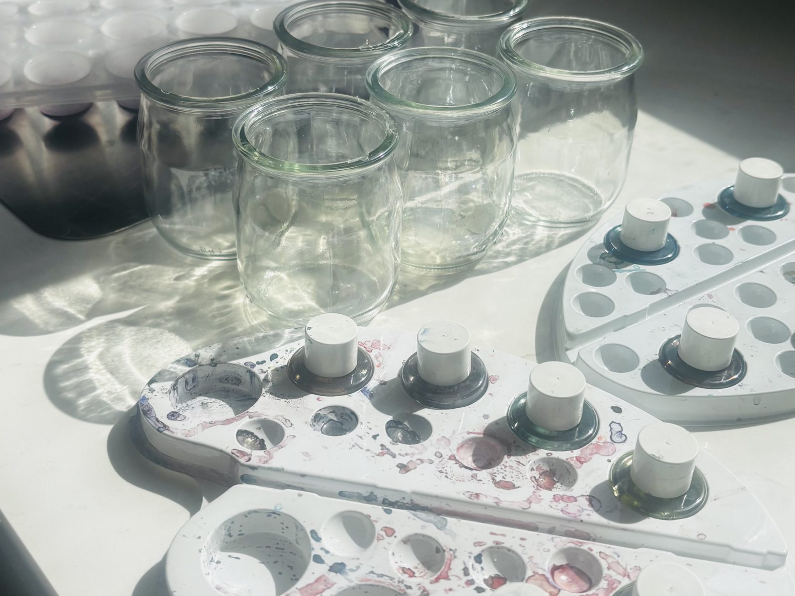 Pots de yaourt en verre et palettes d'aquarelle tachées avec leurs godets d'encre, posés en pleine lumière sur une table blanche