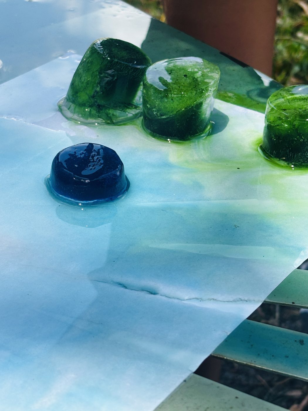 Gros plan sur des glaçons de peinture verts et bleu foncé en train de fondre sur une feuille, la couleur se répand tout autour