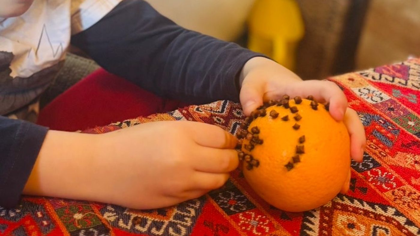 Les mains d'un enfant enfoncent un clou de girofle dans une orange posée sur une nappe colorée, un motif de clous se dessine déjà sur la peau du fruit